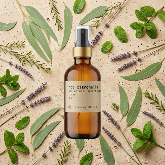 Aire Candle Co. Not Citronella Essential Oil Scented Bug Spray Surrounded by Scent Notes of Lemon Eucalyptus, Tea Tree, Peppermint and Lavender Essential Oils on a Travertine Surface.
