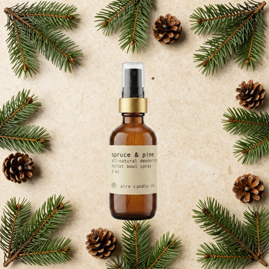 Aire Candle Co. Spruce & Pine Essential Oil Scented Deodorizing Toilet Bowl Spray Surrounded by Scent Notes of Spruce Needles and Pine Cones on a Travertine Surface.
