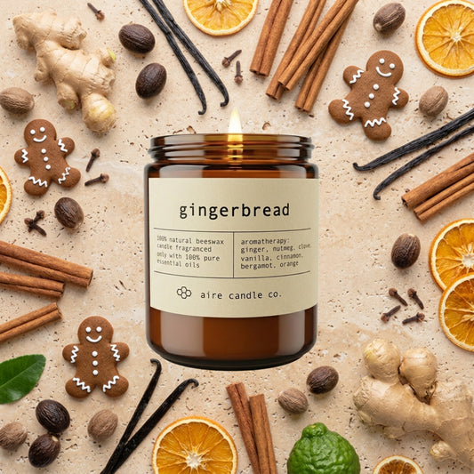 Gingerbread candle from aire candle co surrounded by ginger, gingerbread men, cinnamon, nutmeg, cloves, vanilla beans, bergamots and orange slices on a light travertine background.