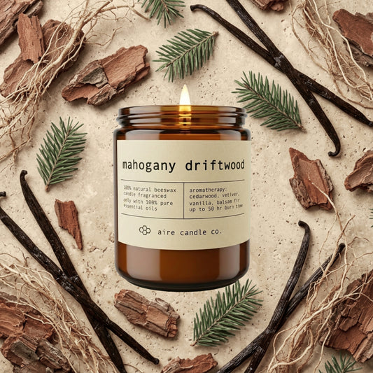 mahogany driftwood beeswax candle from aire candle co surrounded by cedarwood bark, vanilla beans, balsam fir and vetiver on a light travertine background.