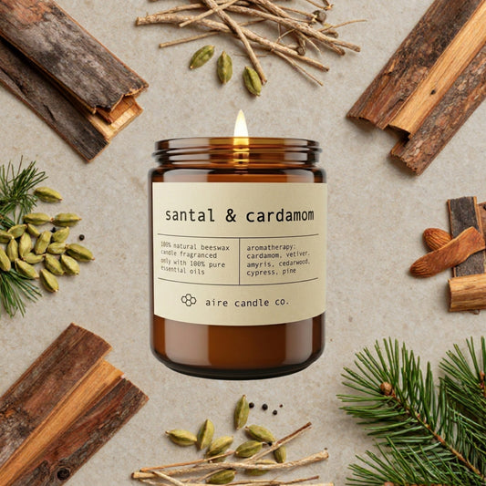 santal & cardamom essential oil scented beeswax candle from aire candle co surrounded by pine needles, cedarwood bark, cypress, cardamom seeds, and vetiver on a light travertine background.