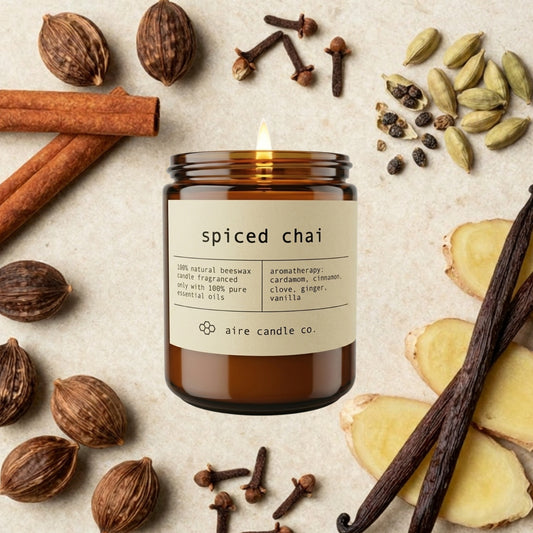 Spiced chai essential oil scented beeswax candle from aire candle co surrounded by cinnamon, ginger, nutmeg, clove, cardamom and vanilla on a light travertine background.