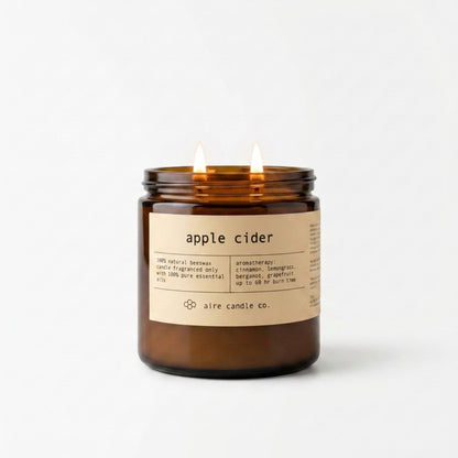 Apple cider candle by aire candle co. in an amber jar with a label on a white background featuring two lit wicks in the 16 oz size.