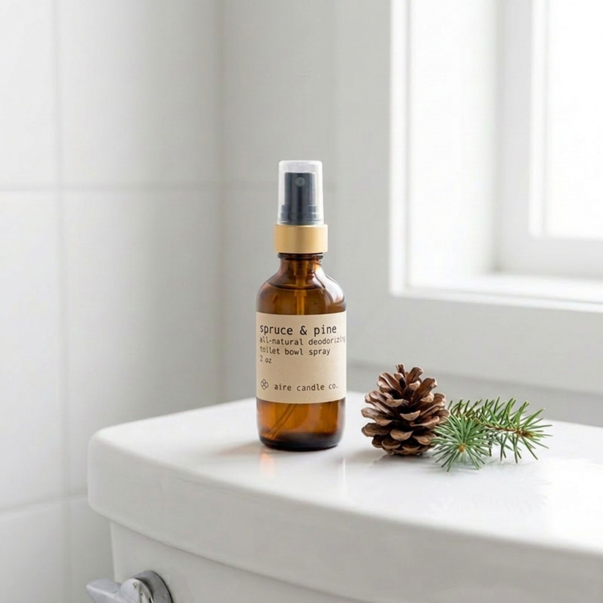 Aire Candle Co. Spruce & Pine Essential Oil Scented Deodorizing Toilet Bowl Spray Sitting Atop a White Toilet Next to a Pine Cone and Spruce Clipping in a Clean, Minimalistic Bathroom Setting