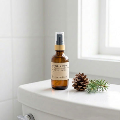 Aire Candle Co. Spruce & Pine Essential Oil Scented Deodorizing Toilet Bowl Spray Sitting Atop a White Toilet Next to a Pine Cone and Spruce Clipping in a Clean, Minimalistic Bathroom Setting