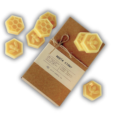 A kraft paper wax melts package tied with twine, labeled 'apple cider,' surrounded by several yellow hexagonal beeswax melts with embossed bee and honeycomb designs on a white background.