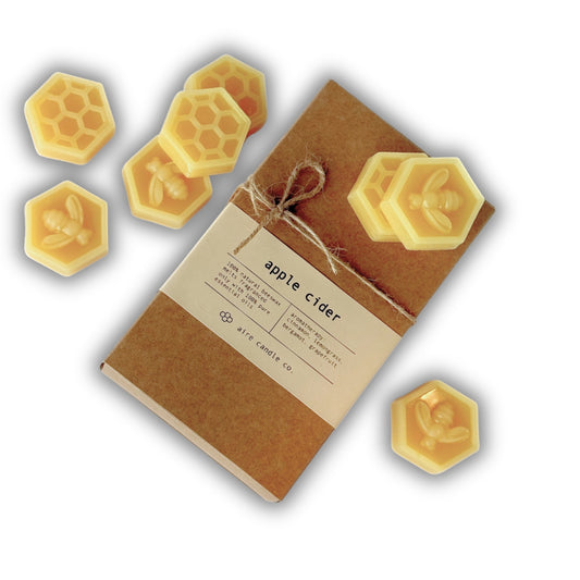 A kraft paper wax melts package tied with twine, labeled 'apple cider,' surrounded by several yellow hexagonal beeswax melts with embossed bee and honeycomb designs on a white background.