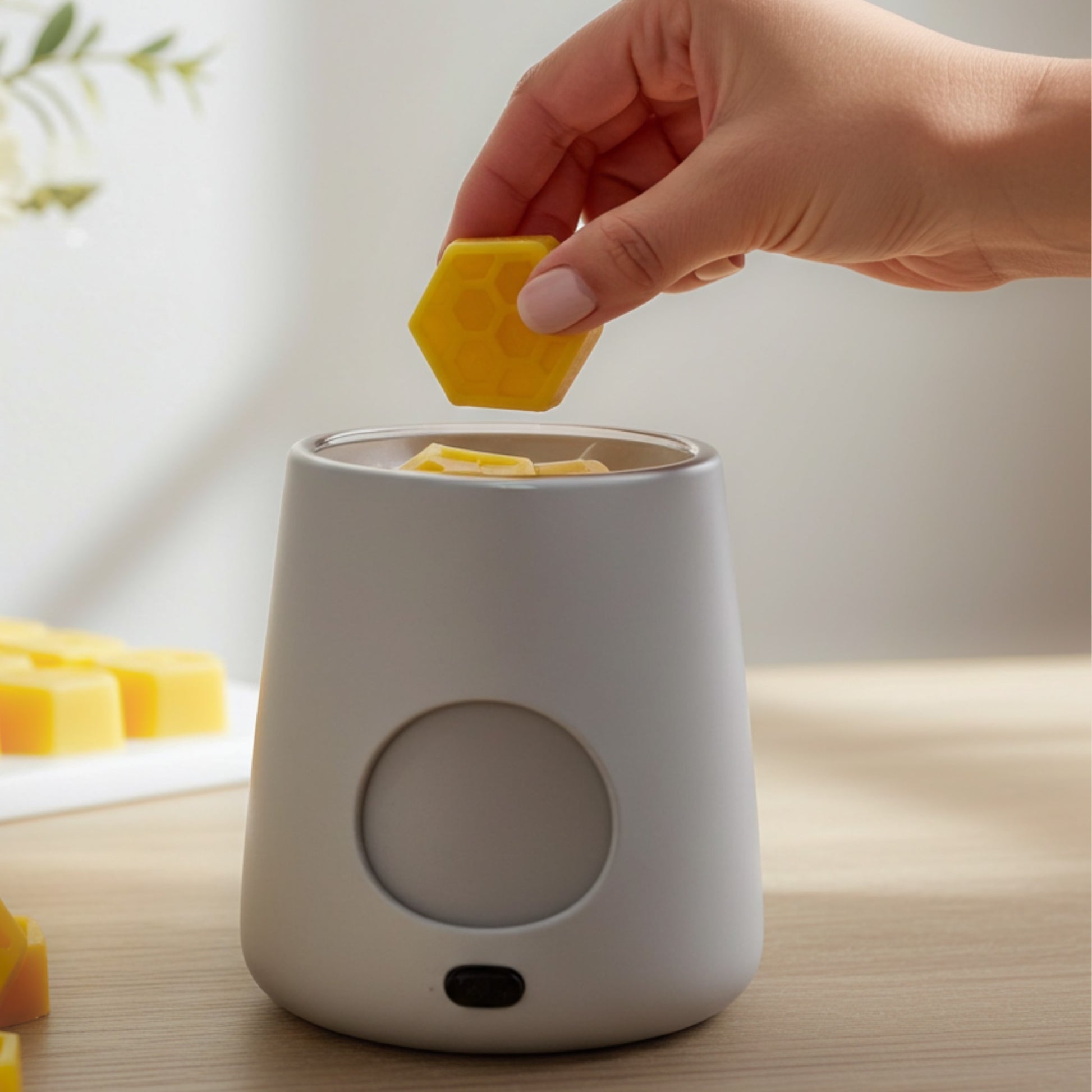 Person placing a yellow honeycomb wax melt into a white electronic wax melter on a light surface.