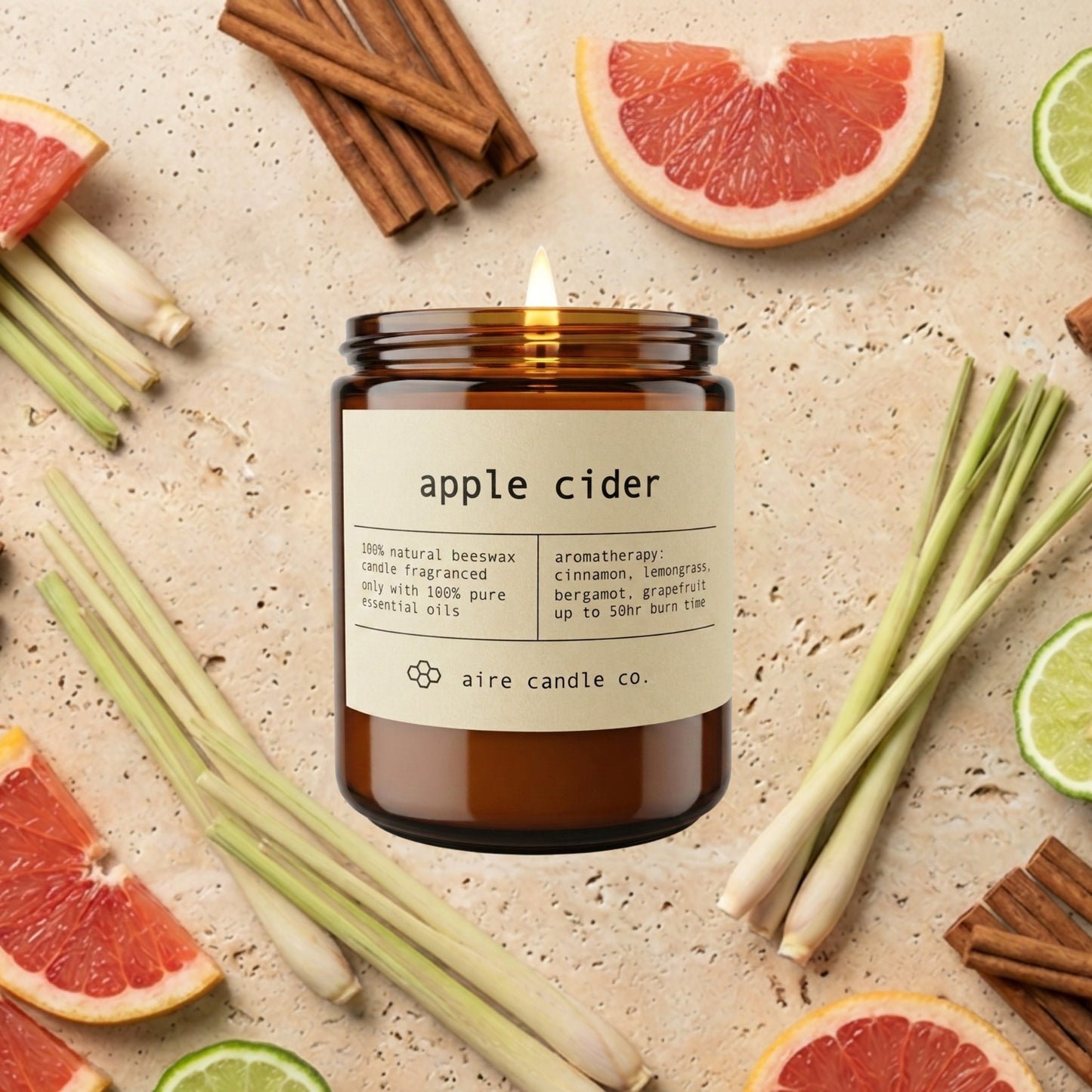 An amber glass candle jar labeled 'apple cider' by aire candle co., styled on a light stone surface with sliced grapefruit, cinnamon sticks, lemongrass, and bergamot wedges around it.