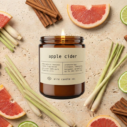 An amber glass candle jar labeled 'apple cider' by aire candle co., styled on a light stone surface with sliced grapefruit, cinnamon sticks, lemongrass, and bergamot wedges around it.