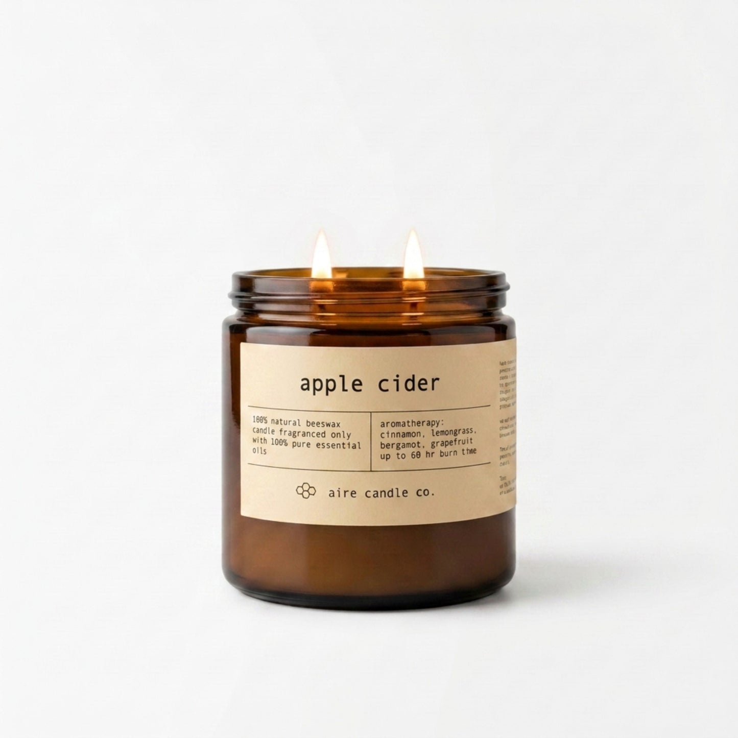 Apple cider candle by aire candle co. in an amber jar with a label on a white background featuring two lit wicks in the 16 oz size.