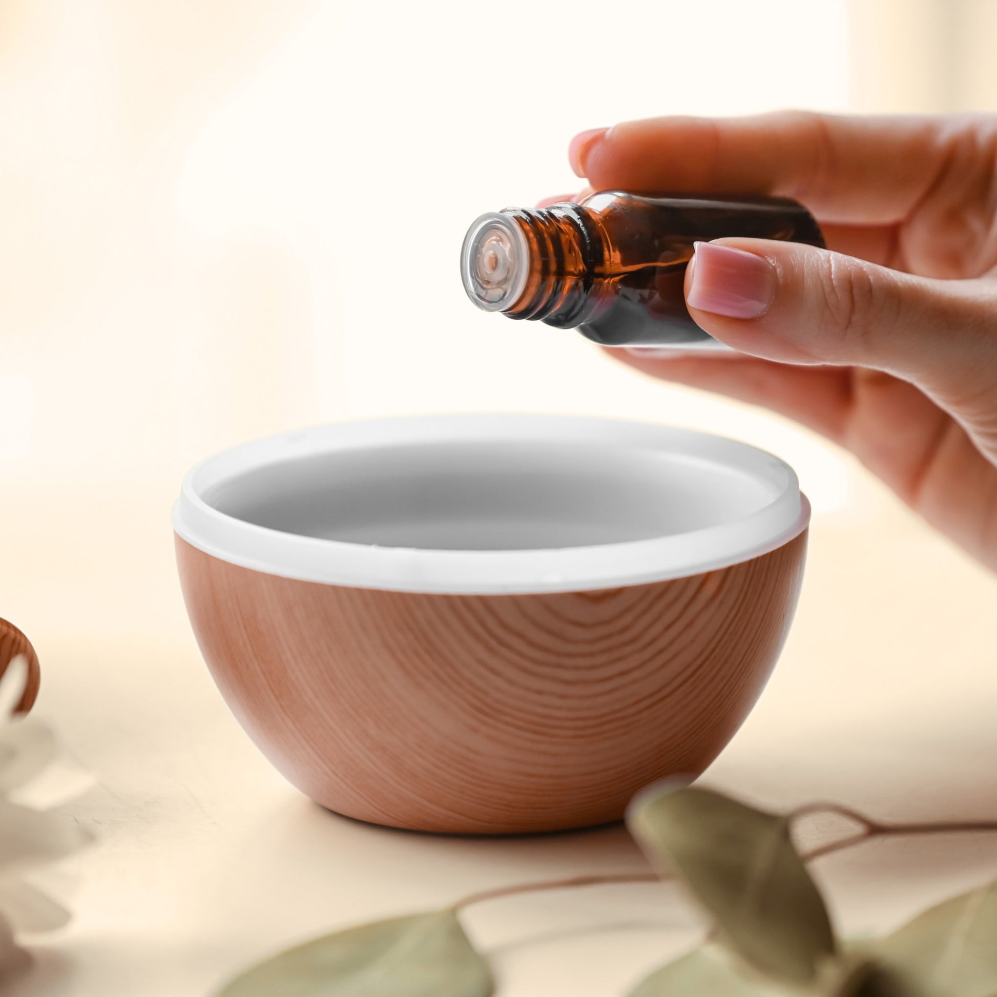 Hand holding a small amber essential oil bottle over a wooden essential oil diffuser bowl with a white interior.