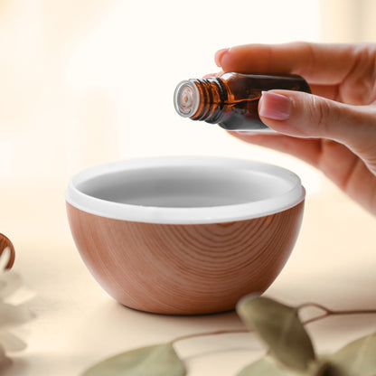 Hand holding a small amber essential oil bottle over a wooden essential oil diffuser bowl with a white interior.