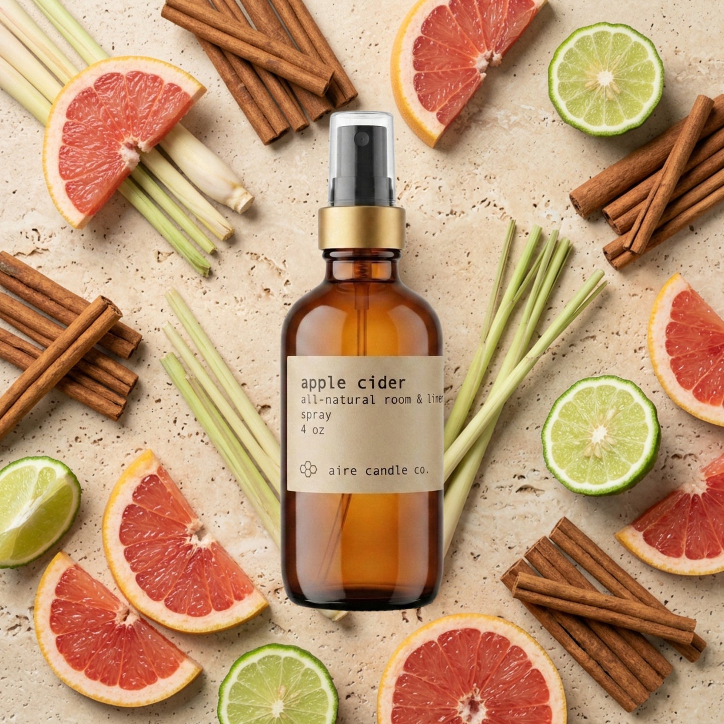 All-natural apple cider scented room spray featuring a black fine mist spray head, gold rim and amber glass bottle with a kraft paper label. The background shows grapefruit slices, cinnamon, lemongrass and bergamot fruits against a beige travertine surface.