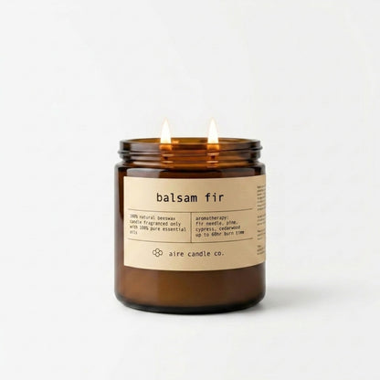 16 oz balsam fir candle by aire candle co. in an amber jar showing two wicks illuminated and a label on a white background