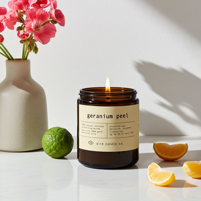 A lit beeswax candle labeled 'geranium peel' with a vase of pink flowers, a lime, and clementine slices on a white surface.