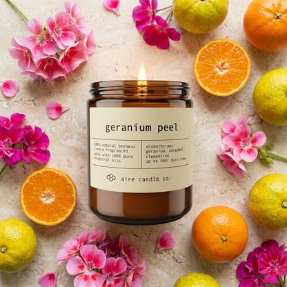 Geranium Peel candle from aire candle co surrounded by geranium flowers, clementines and bergamots on a light travertine background.