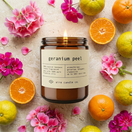 Geranium Peel candle from aire candle co surrounded by geranium flowers, clementines and bergamots on a light travertine background.