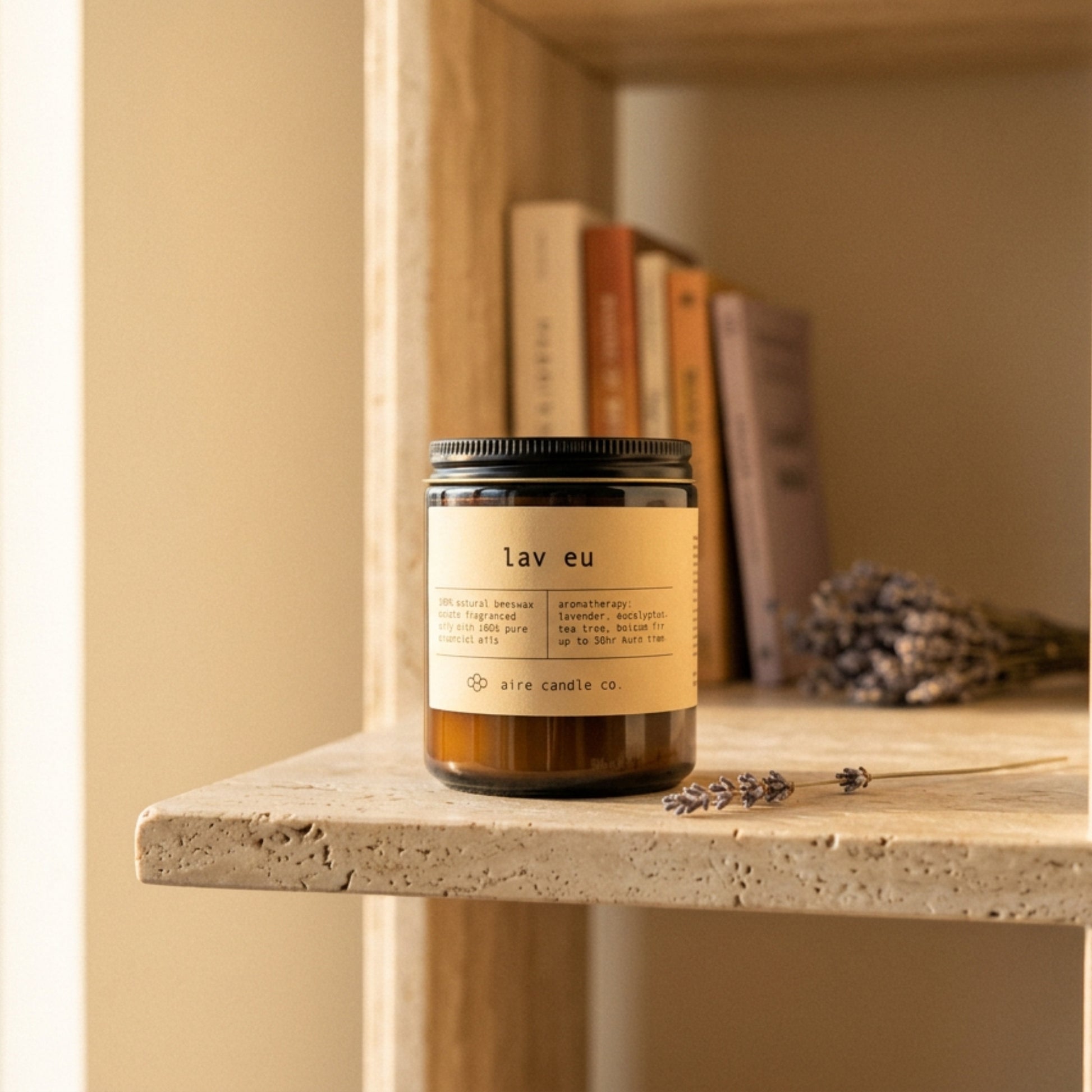 Candle labeled 'lav eu' by aire candle co. on a travertine shelf with books and lavender sprigs.