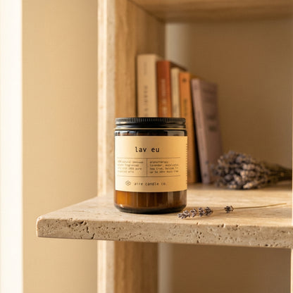 Candle labeled 'lav eu' by aire candle co. on a travertine shelf with books and lavender sprigs.
