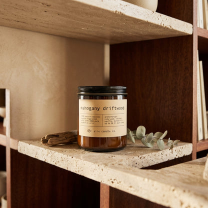 Candle labeled 'mahogany driftwood' by aire candle co. on a travertine shelf with books and decorative items.