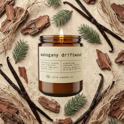mahogany driftwood beeswax candle from aire candle co surrounded by cedarwood bark, vanilla beans, balsam fir and vetiver on a light travertine background.