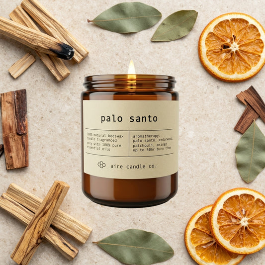 palo santo essential oil scented beeswax candle from aire candle co surrounded by palo santo sticks, dried oranges, patchouli leaves and cedarwood on a light travertine background.