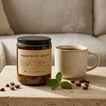 Beeswax candle labeled 'peppermint mocha' by aire candle co. on a travertine surface with a steaming coffee mug and coffee beans.
