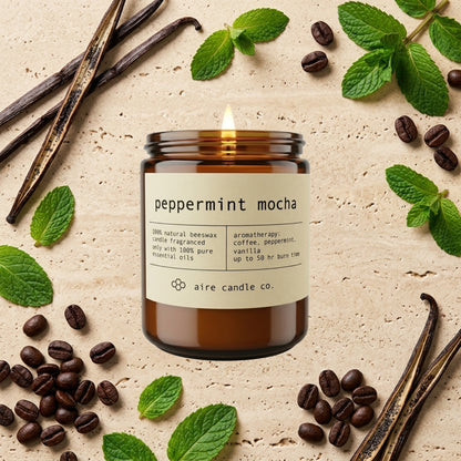 peppermint mocha essential oil scented beeswax candle from aire candle co surrounded by peppermint leaves, coffee beans and vanilla beans on a light travertine background.