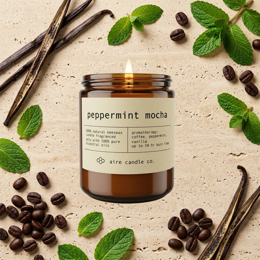 peppermint mocha essential oil scented beeswax candle from aire candle co surrounded by peppermint leaves, coffee beans and vanilla beans on a light travertine background.