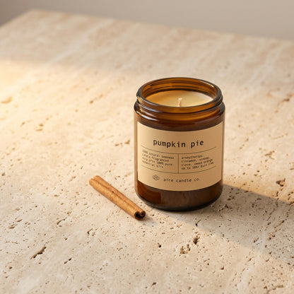 Amber glass candle labeled 'pumpkin pie' with a cinnamon stick on a stone surface