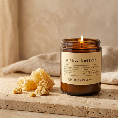 Purely beeswax candle in an amber jar with a label, surrounded by honeycomb and cloth on a travertine surface.