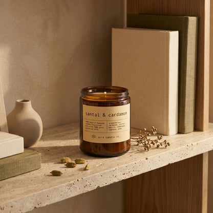 Scented candle labeled 'santal & cardamom' on a shelf with books and a vase.