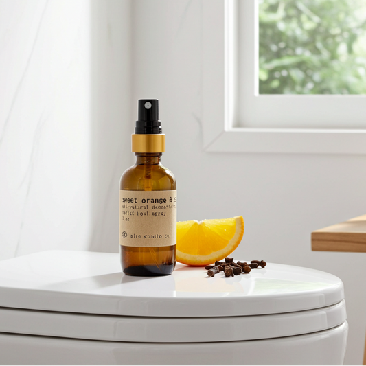 Bottle of essential oil spray on a toilet seat with an orange and cloves in a bathroom.