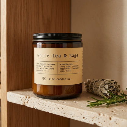 Beeswax candle labeled 'white tea & sage' by aire candle co. on a travertine shelf with books and decorative items.