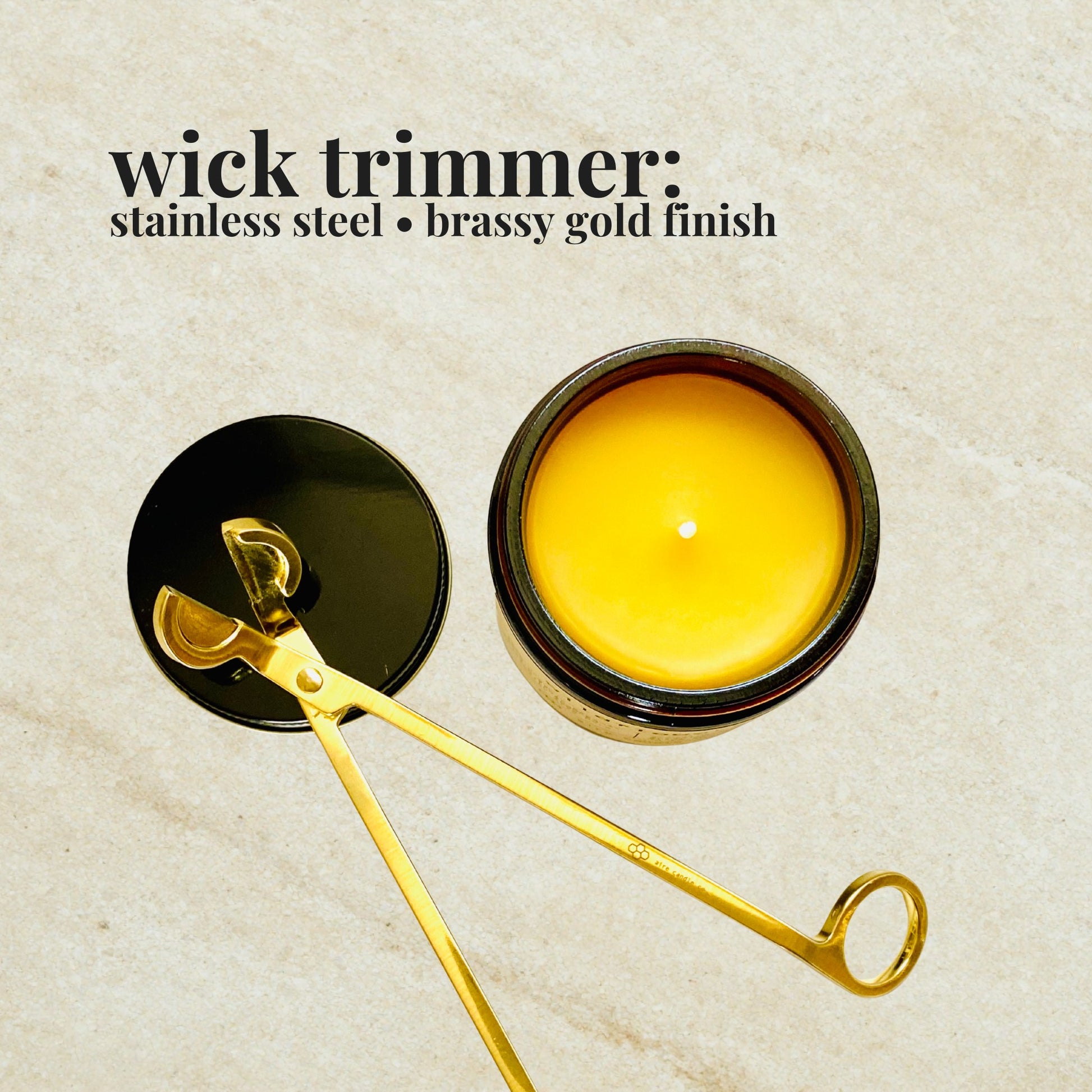 Gold wick trimmer resting on a black metal lid, next to a yellow beeswax candle on a beige background.