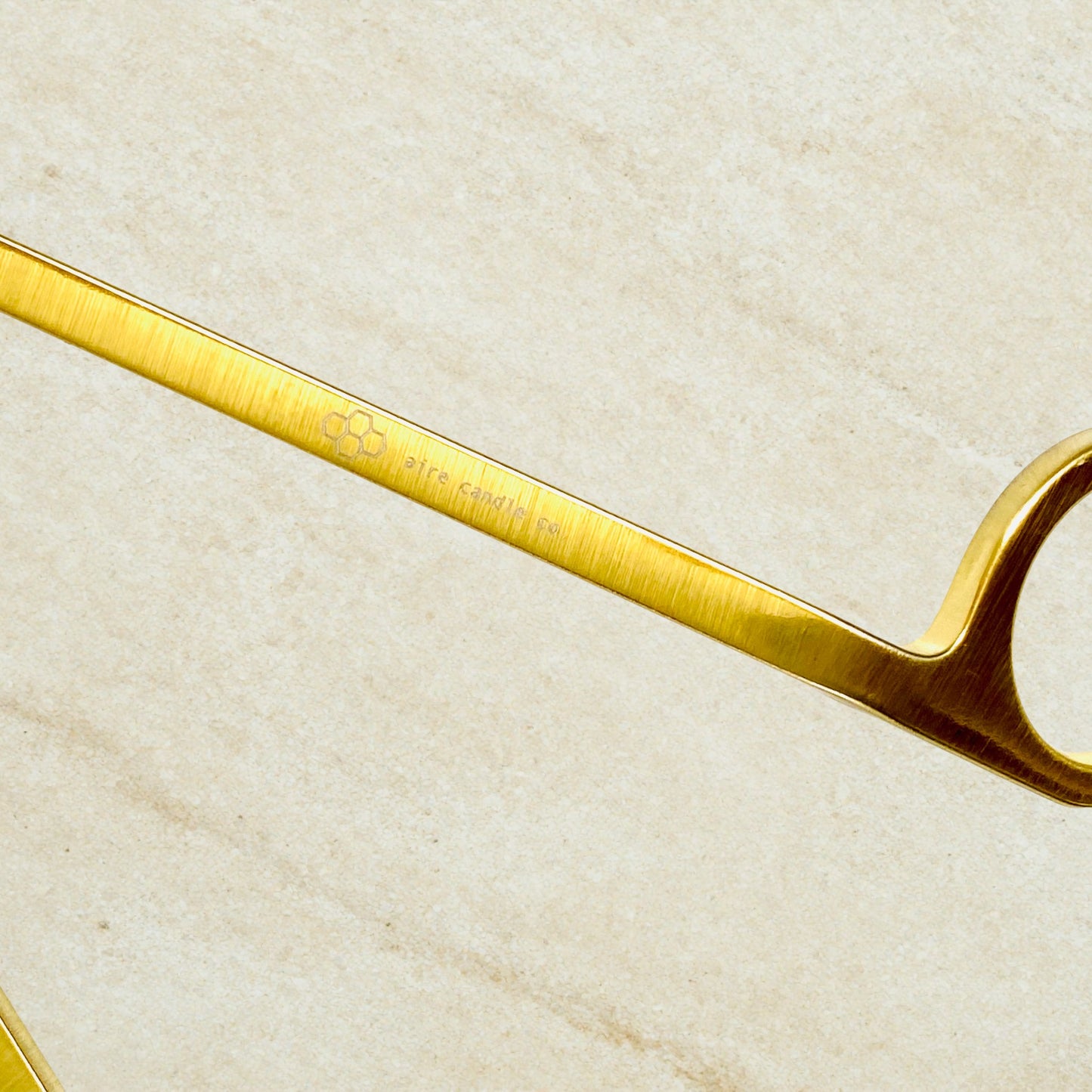 Gold wick trimmer on a beige textured background showcasing the aire candle co. engraving on the wick trimmer's handle.