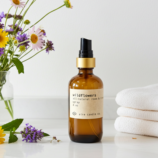 A bottle of 'Wildflowers' room spray by aire candle co. on a white surface with flowers and towels in the background.